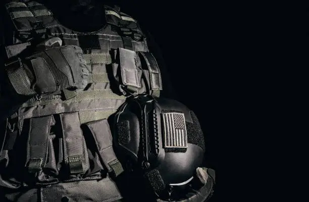 a soldier in tactical gloves and armor vest standing and holding military helmet with american flag patch on black background.