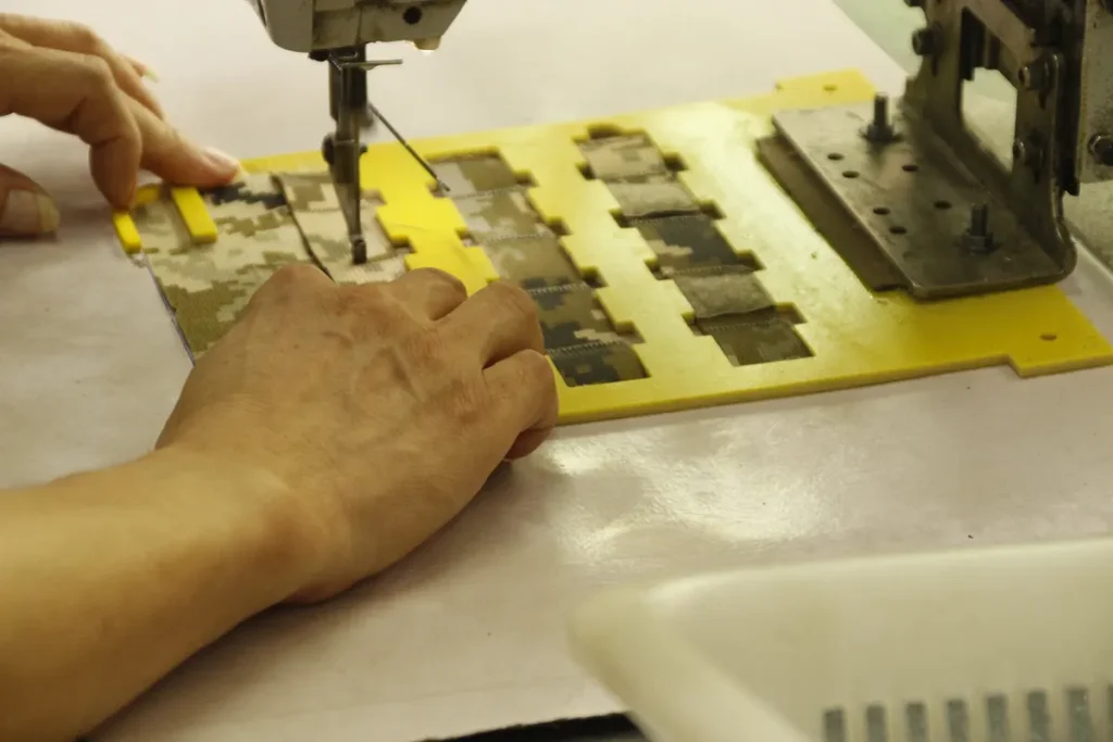 A worker sewing camouflage fabric pieces on a yellow cutting mat using an industrial sewing machine in a tactical gear manufacturing setting.