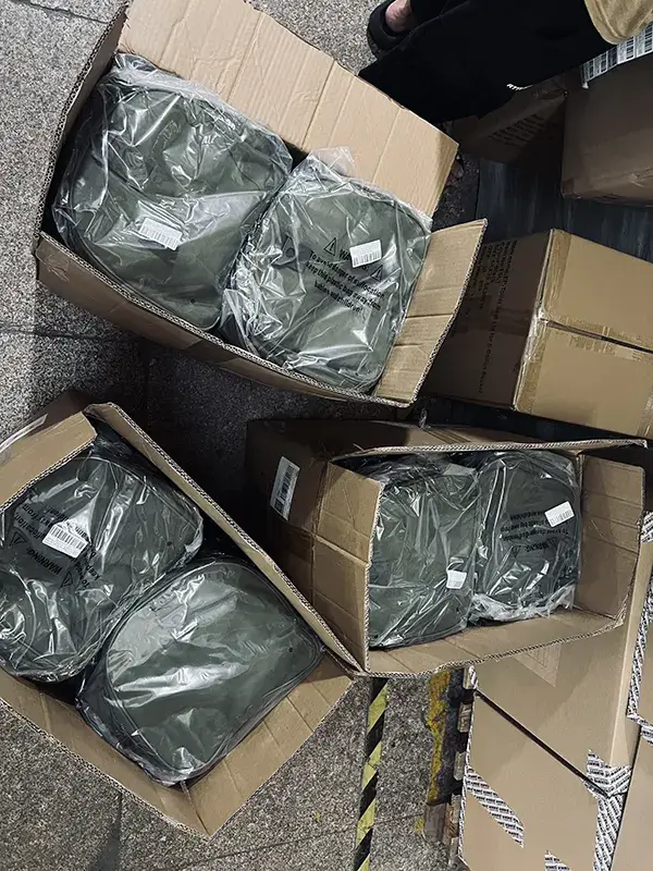 Open cardboard boxes containing green tactical gear bags wrapped in plastic, stacked on our warehouse floor